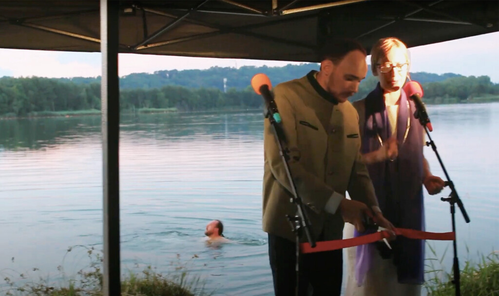 Zwei Personen stehen vor Mikrofonen, hinter ihnen ein Badesee